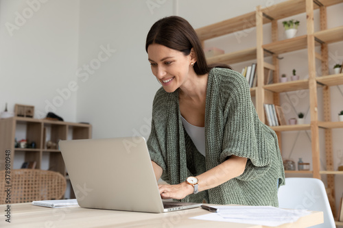 Interested smiling young businesswoman standing near table in modern office, using laptop, positive female employee looking at screen, writing email, chatting, working on online project © fizkes