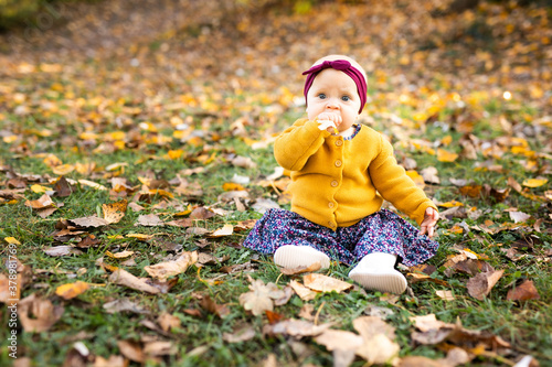 Baby girl in yelloy jacket and red headband seating on the grass, playing in the autumn leaves