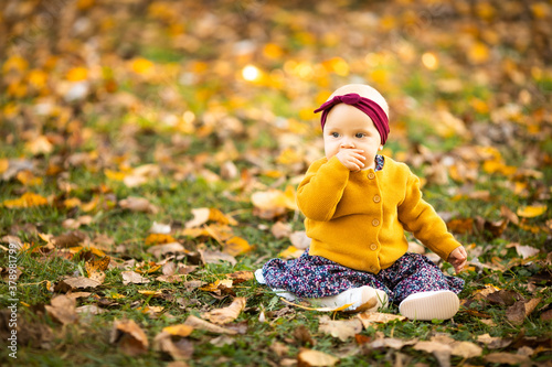 Baby girl in yelloy jacket and red headband seating on the grass  playing in the autumn leaves
