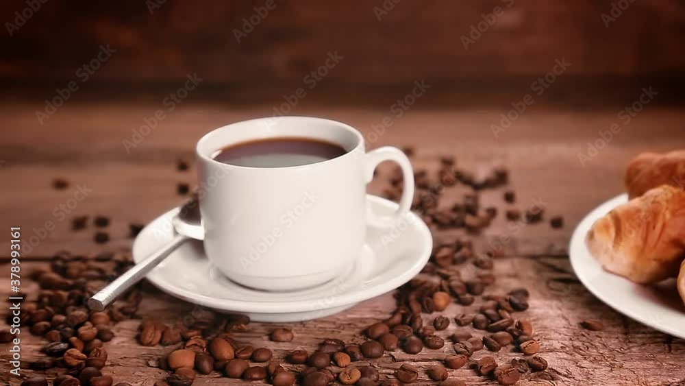 A woman's hand places a plate of croissants next to a cup of coffee and coffee beans scattered on the table. A woman's hand places a plate of croissants next to a cup of coffee and coffee beans scattered on the table.