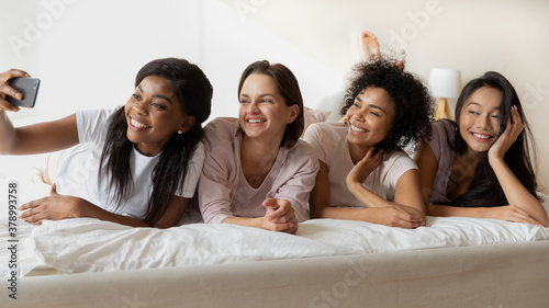 Head shot happy attractive young african american girl holding smartphone, taking selfie with smiling pretty multiracial friends lying together on bed, entertaining together at hen sleepover party.