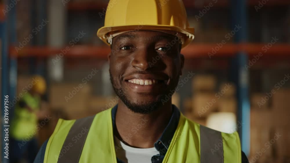 Handsome and Happy Professional Worker Wearing Safety Vest and Hard Hat Charmingly Smiling on Camera. In the Background Big Warehouse with Shelves full of Delivery Goods. Medium Close-up Portrait Handsome and Happy Professional Worker Wearing Safety Vest and Hard Hat Charmingly Smiling on Camera. In the Background Big Warehouse with Shelves full of Delivery Goods. Medium Close-up Portrait