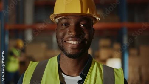 Handsome and Happy Professional Worker Wearing Safety Vest and Hard Hat Charmingly Smiling on Camera. In the Background Big Warehouse with Shelves full of Delivery Goods. Medium Close-up Portrait photo