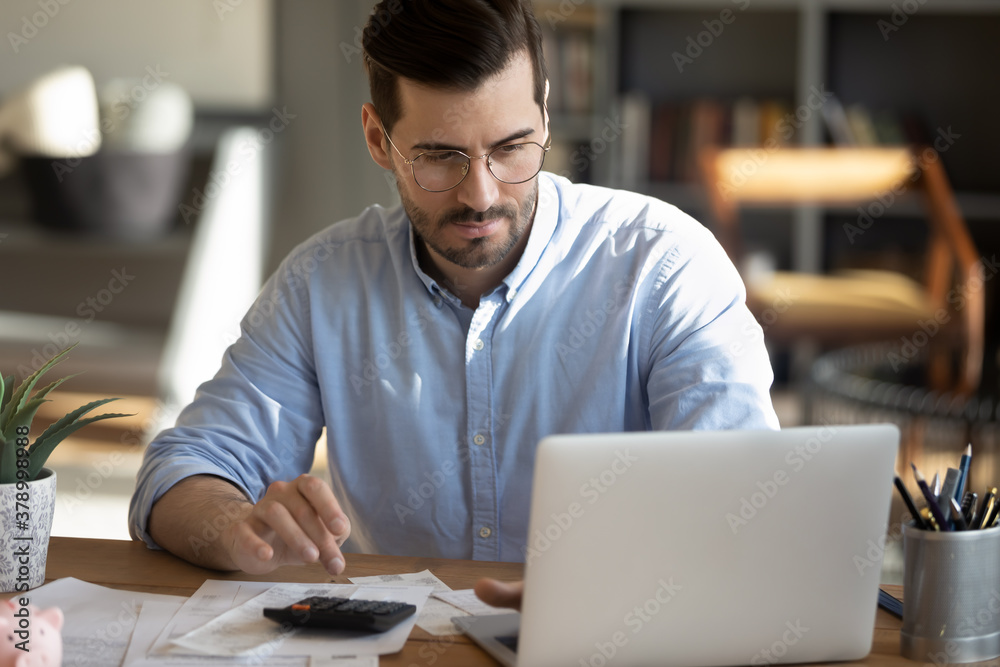 Focused young Caucasian man look at laptop screen calculate expenses expenditures pay bills taxes online. Millennial male busy managing household family budget, take care of financial paperwork. Focused young Caucasian man look at laptop screen calculate expenses expenditures pay bills taxes online. Millennial male busy managing household family budget, take care of financial paperwork.