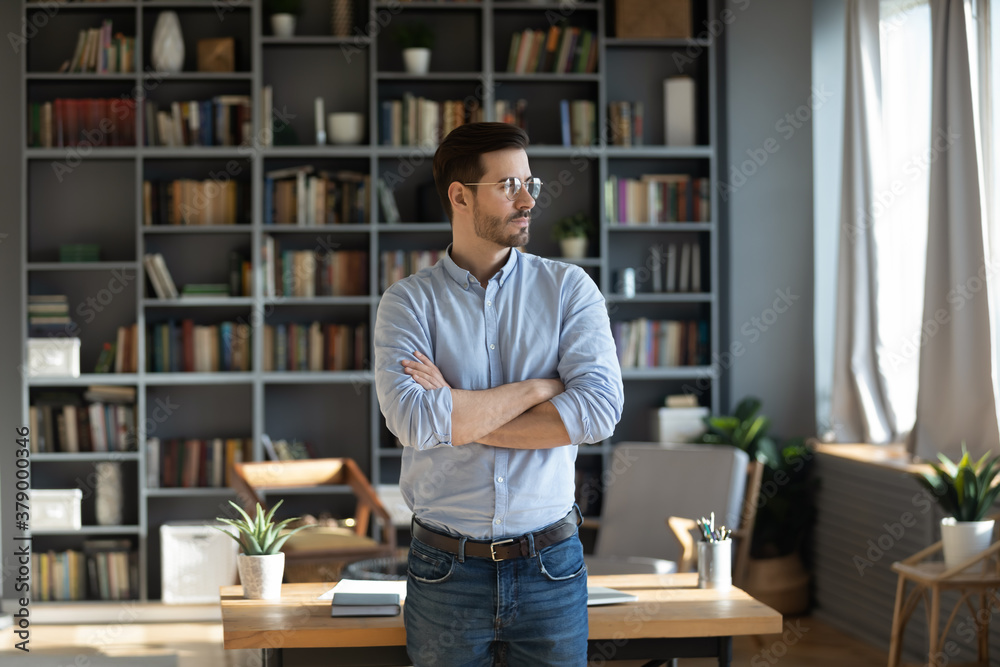 Obraz premium Serious confident young 30s Caucasian businessman stand near home office desk look in distance thinking or planning. Pensive millennial male pondering making decisions. Business vision concept.