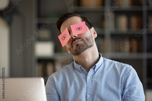 Exhausted young Caucasian man worker have sticker pads on eyes sleeping near computer in office. Tired millennial male employee fall asleep doze off at workplace, feel overwhelmed drained at work.