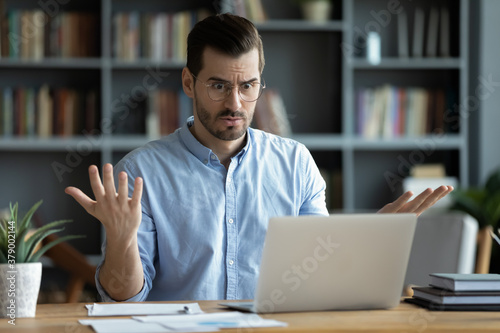 Unhappy young Caucasian male worker in glasses look at laptop screen shocked by gadget breakdown or operational problems. Frustrated man confused surprised by unexpected error on computer device.
