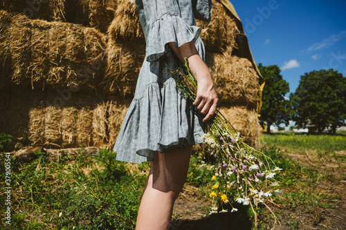 Young girl in rubber boots with flowers standing against the background of straw bales on country farm. Farming, Cottagecore, Farmcore, Countrycore aesthetics, fresh air, countryside, slow life photo