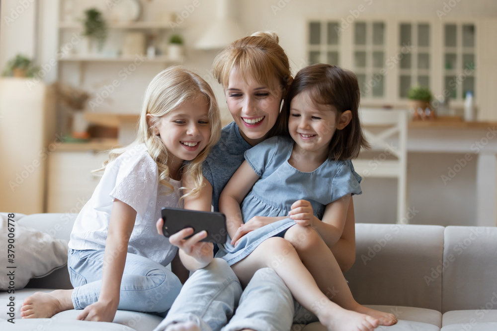 Happy mom and two daughters watching funny video on smartphone sitting on comfortable couch in cozy living room. Smiling mother and children enjoying cartoons on mobile phone at weekend Happy mom and two daughters watching funny video on smartphone sitting on comfortable couch in cozy living room. Smiling mother and children enjoying cartoons on mobile phone at weekend