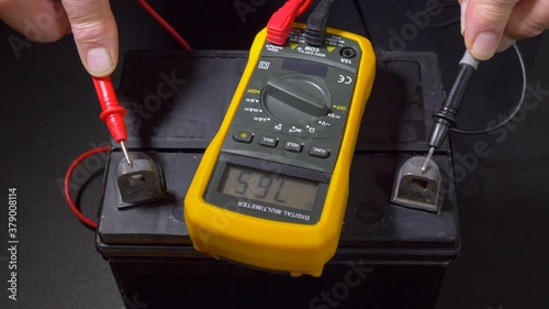 Man switching on a multimeter on a vehicle battery, then connecting red and black probes to the positive and negative terminals, to confirm from the low voltage reading that the battery is discharged. photo