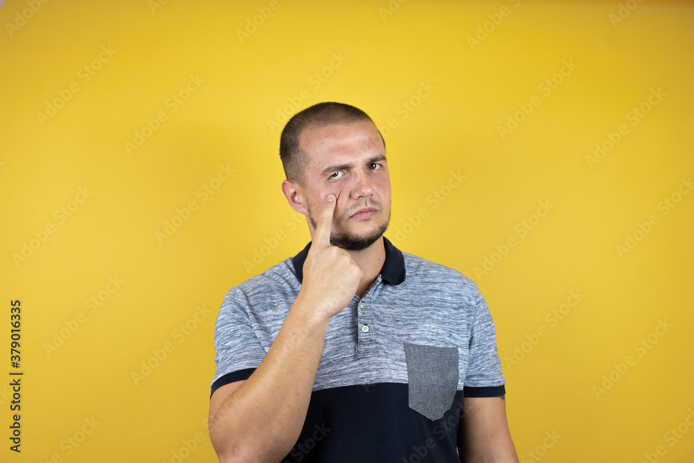 Russian man standing over insolated yellow background Pointing to the eye watching you gesture, suspicious expression Russian man standing over insolated yellow background Pointing to the eye watching you gesture, suspicious expression