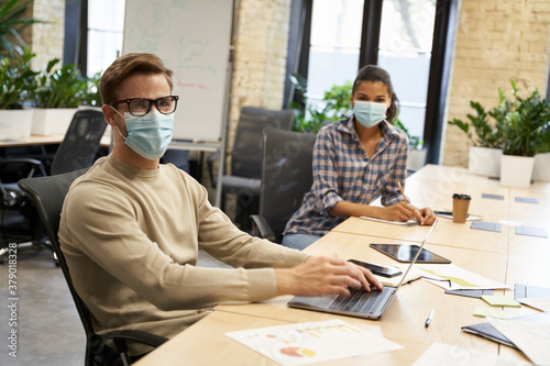 Working safely. Male and female office workers wearing medical protective masks sitting together at the table in the modern office and looking at camera