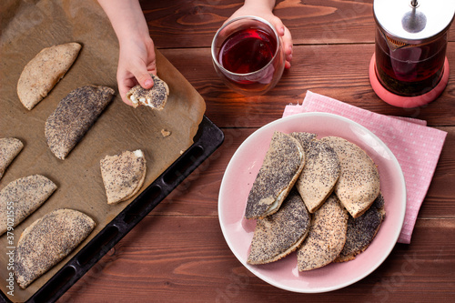 Child hands holding traditional Russian sweet shortcrust pastry with quark or cottage cheese and raisins, sochni or sochniki pies or pirozhki on pink plate with tea in the morning on wooden table photo