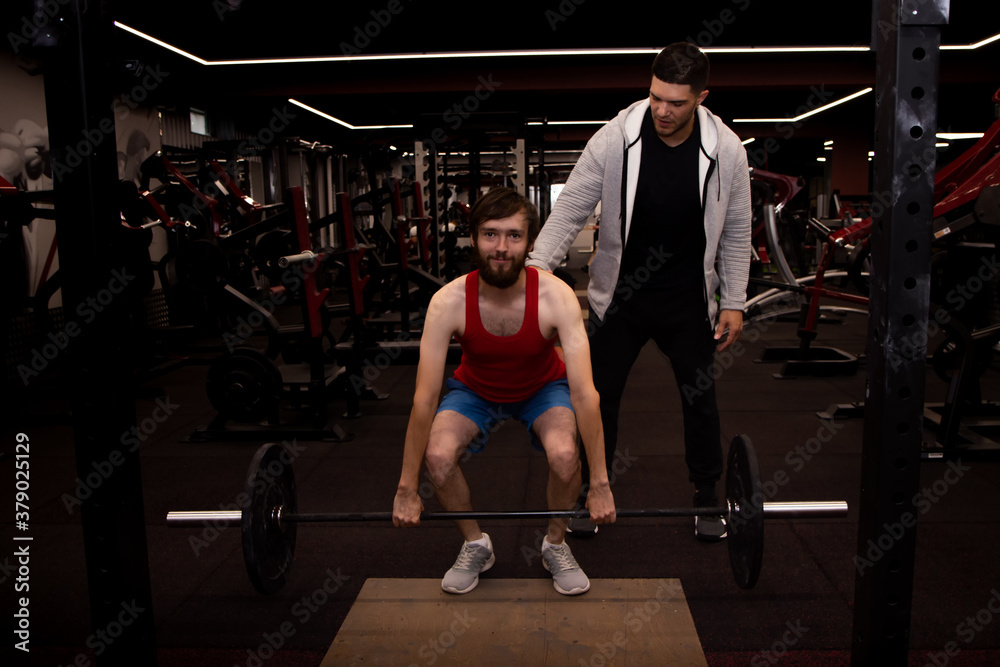 Young man doing exercises with a personal trainer as he lifts a barbell weight to chest height in gym Young man doing exercises with a personal trainer as he lifts a barbell weight to chest height in gym