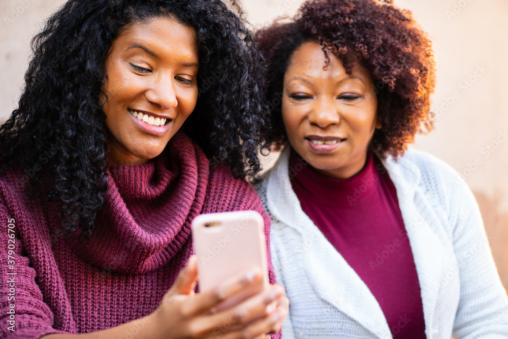 Cheerful latin adult daughter and mother with smart cell phone looking at cell phone screen outside the house. Family, affectionate, reunion, together concept.. - obrazy, fototapety, plakaty 