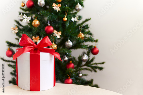 Christmas present box on table near Christmas tree. White round gift box with red ribbon against decorated christmas tree background. Concept of Christmas holidays