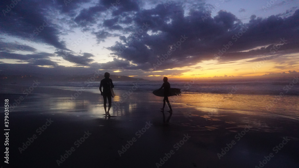 Silueta de dos amigos caminando con tabla de surf en la orilla de la playa con la última luz del atardecer en el horizonte en un día con sol, nubes y montañas con pueblo al fondo