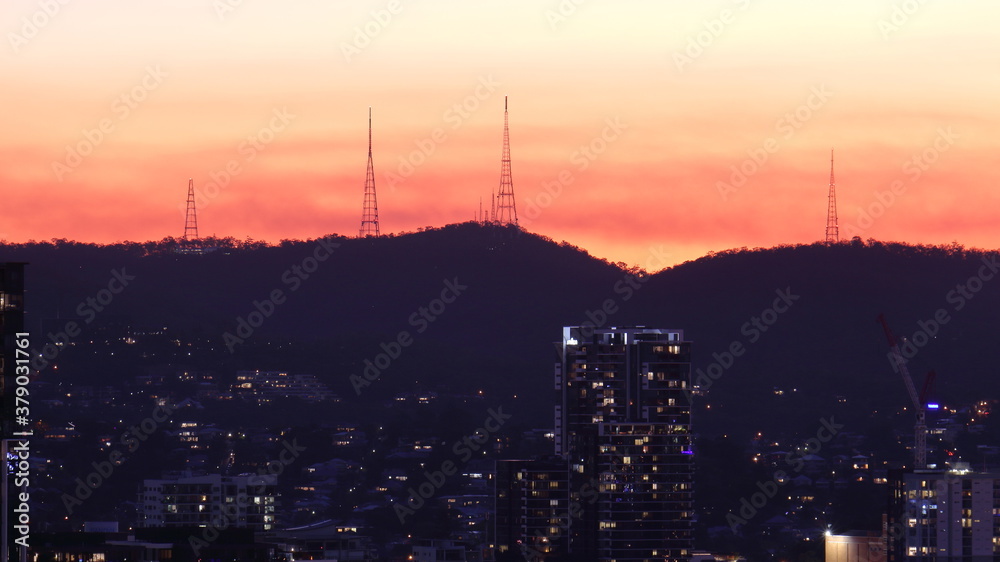 Silhouette of radio and tv transmission towers against the setting sun, on Mount Coot-Tha, with buildings in the foreground, in Brisbane, Queensland, Australia