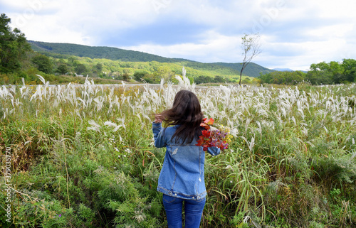 Young woman holding bouquet of autumn wildflowers and branches with red leaves, standing with her back to the camera and looking at field of Miscanthus sinensis (Chinese silver grass). Touching hair