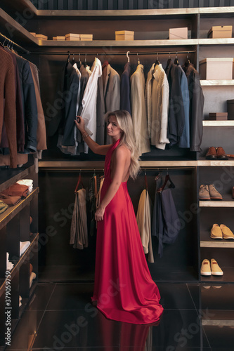Elegant caucasian woman in red long train dress stands in dark closet full of menswear. Fashion theme.