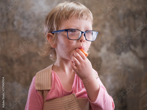 funny little blond boy in pink sweatshirt, big glasses and corset to correct posture eating fresh carrot. concept of vegetarianism and proper balanced nutrition for children