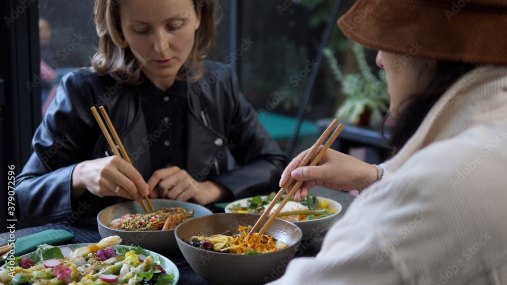 A girl in a hat feeds blonde girl with chopsticks. Sharing your food with the loved ones. 