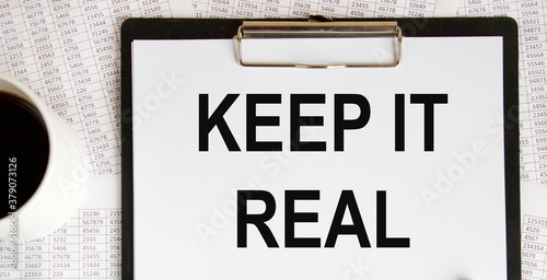 KEEP IT REAL text on paper sheet with chart, dice, spectacles, pen, laptop and blue and yellow push pin on wooden table - business, banking, finance and investment concept