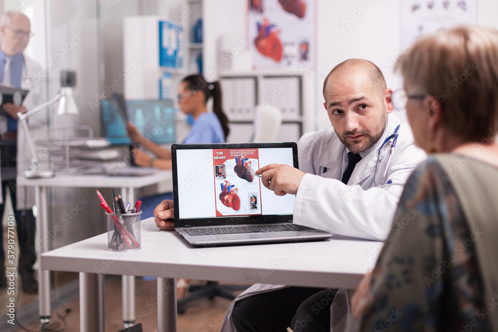 Doctor looking at senior woman while pointing at heart diagram in hospital office during consultation. Nurse in blue uniform holding x-ray image. Doctor looking at senior woman while pointing at heart diagram in hospital office during consultation. Nurse in blue uniform holding x-ray image.