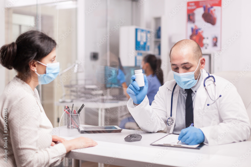 Doctor with face mask against coronavirus outbreak holding pills bottle while writing prescription for sick woman. Nurse wearing blue uniform working on computer. Doctor with face mask against coronavirus outbreak holding pills bottle while writing prescription for sick woman. Nurse wearing blue uniform working on computer.