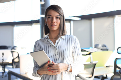 Portrait of young businesswoman holding touch pad while standing in modern office space interior.