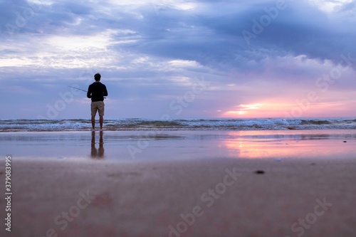 A young man fishing in the open sea along the Atlantic Ocean shoreline at Costa da Caparica in Lisbon, Portugal. Attractive guy holding the fishing rod at sunset on the beach. Hobby lifestyle © Martina