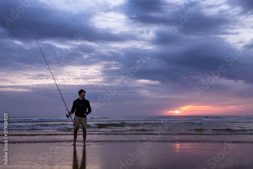 A young man fishing in the open sea along the Atlantic Ocean shoreline at Costa da Caparica in Lisbon, Portugal. Attractive guy holding the fishing rod at sunset on the beach. Hobby lifestyle