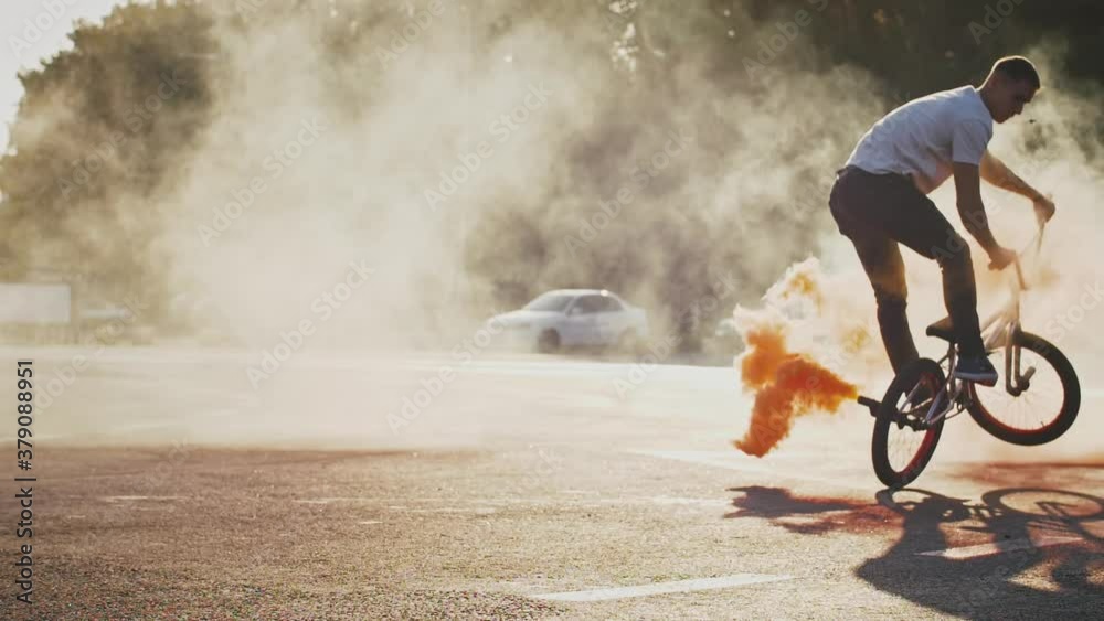 Young male is performing difficult tricks riding bmx bike with orange smoke bomb attached to its rear wheel