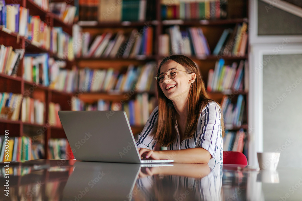 Young smiling attractive college girl sitting in library and using laptop. She is writing homework. Young smiling attractive college girl sitting in library and using laptop. She is writing homework.