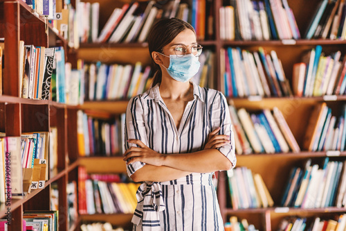 Young smart attractive female student with face mask standing in library with arms crossed. Studying during corona virus pandemic concept.