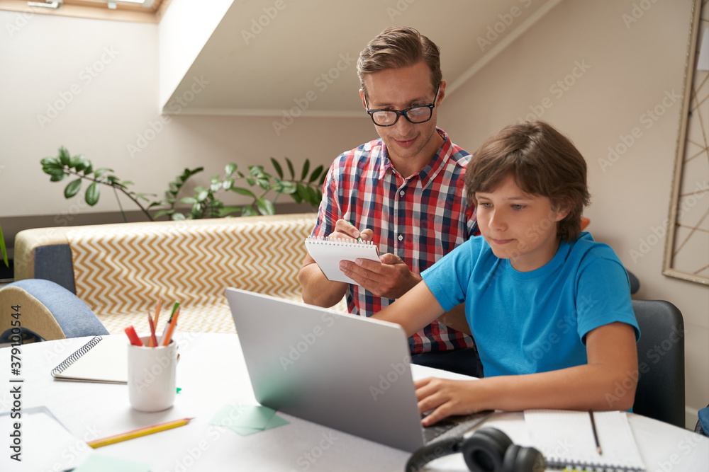 Homeschooling concept. Young father wearing eyeglasses and his little son watching video lesson online, man making some notes Homeschooling concept. Young father wearing eyeglasses and his little son watching video lesson online, man making some notes