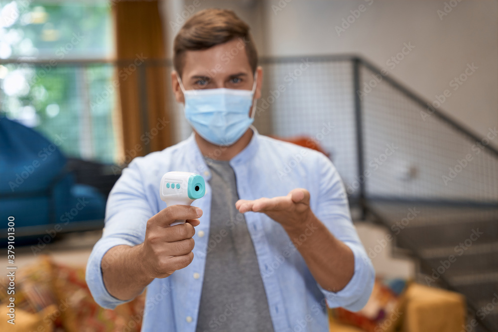 Business during Covid 19 or coronavirus, portrait of young male office worker wearing medical protective mask standing in the office and showing infrared thermometer at camera