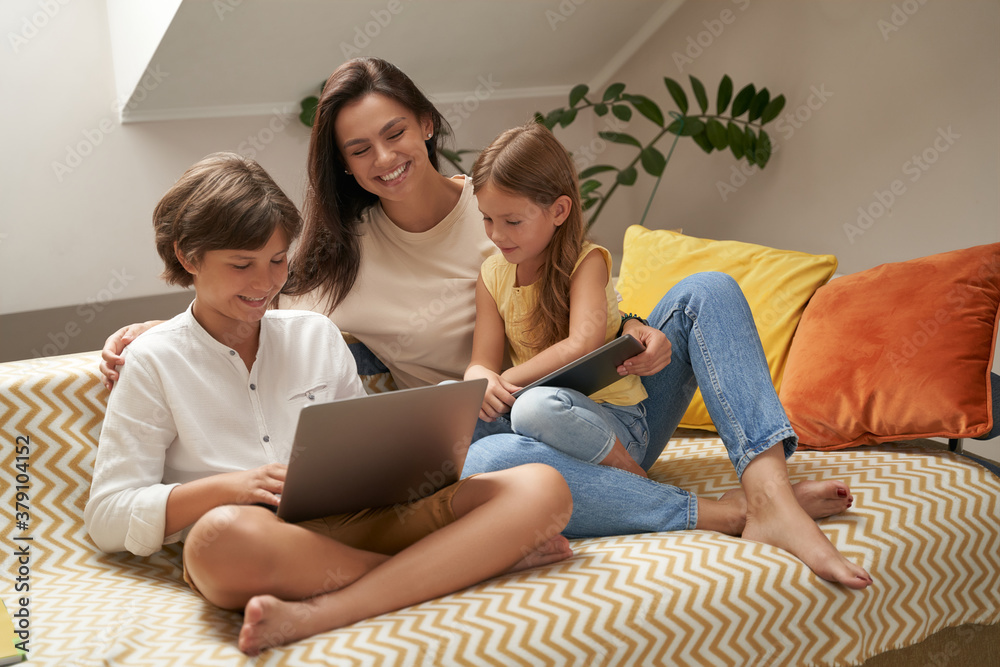 Young beautiful caucasian family, mother and her two cute little kids relaxing on sofa, watching funny videos or cartoons on laptop Young beautiful caucasian family, mother and her two cute little kids relaxing on sofa, watching funny videos or cartoons on laptop