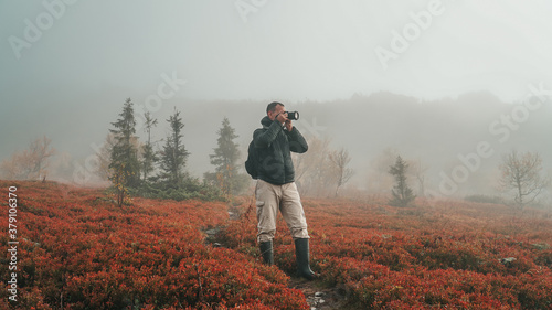 Young Caucasian Male Photographer Taking Picture of Nordic Nature in Early Misty Morning with Fog among Red Bilberry Bushes in Autumn