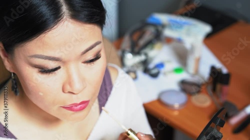 Middle-aged woman puts a concealer on her face and smears it with her fingers. She holds a mirror and applies makeup. High quality 4k footage.