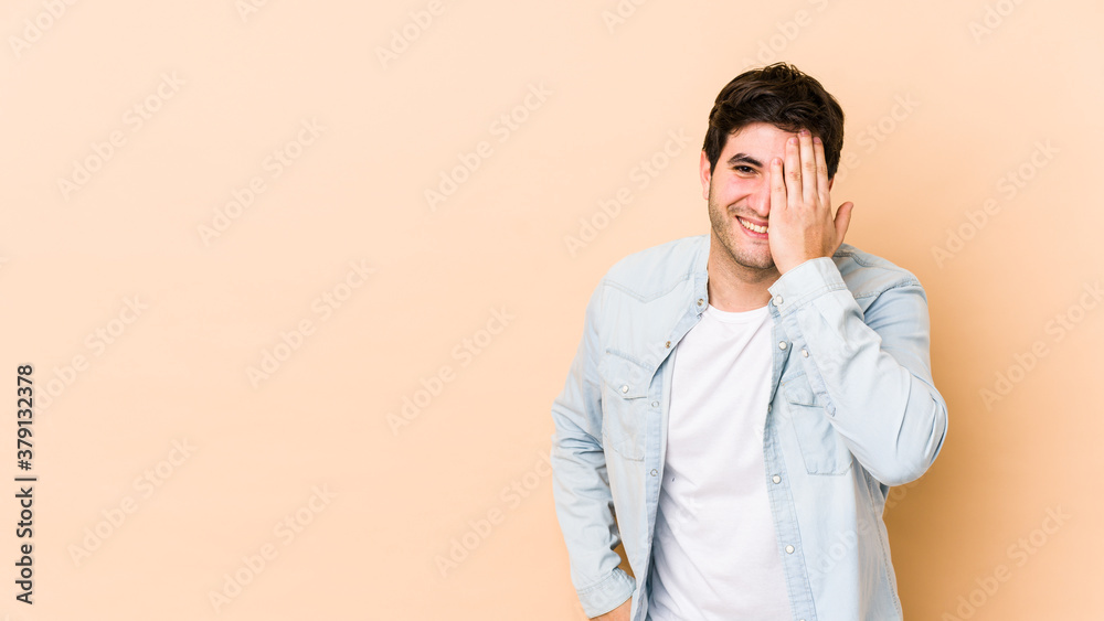 Young man isolated on beige background having fun covering half of face with palm. Young man isolated on beige background having fun covering half of face with palm.