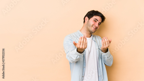 Young man isolated on beige background pointing with finger at you as if inviting come closer.