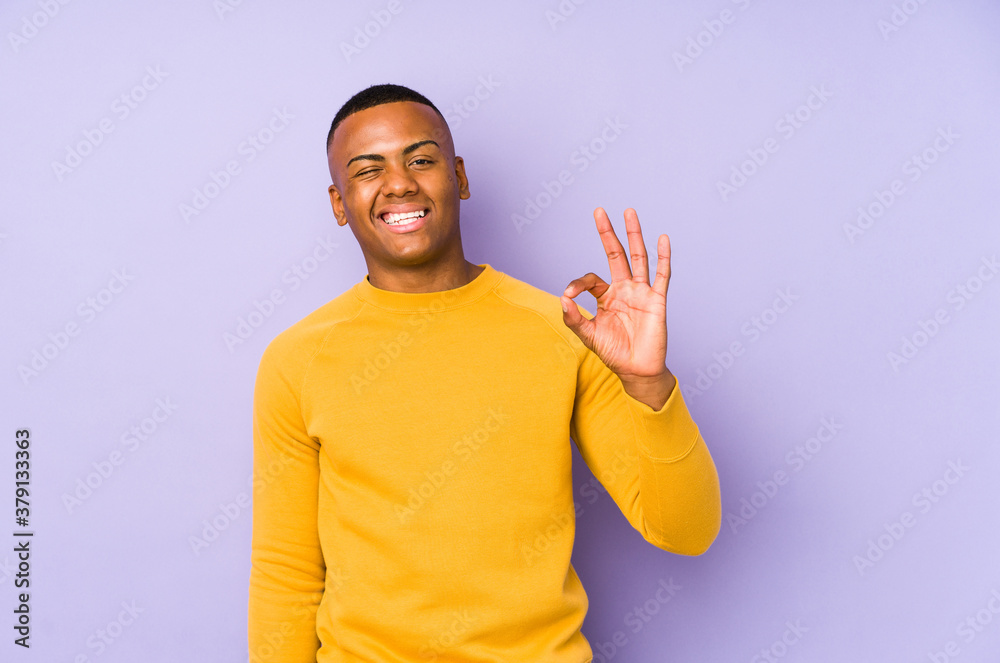 Young latin man isolated on purple background winks an eye and holds an okay gesture with hand. Young latin man isolated on purple background winks an eye and holds an okay gesture with hand.