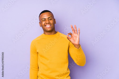Young latin man isolated on purple background winks an eye and holds an okay gesture with hand.