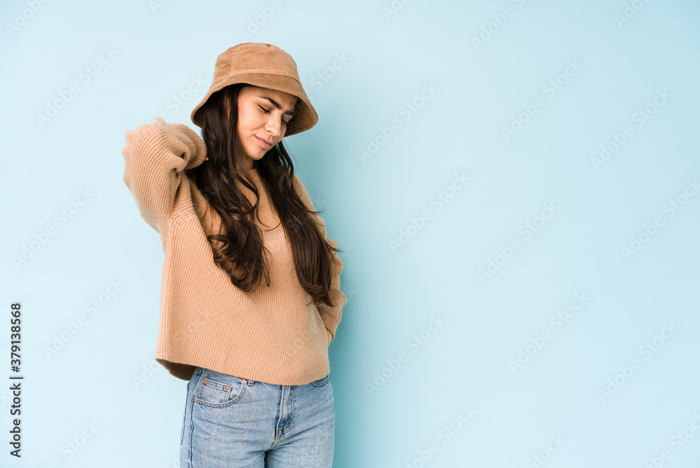 Young indian woman wearing a hat isolated on blue background having a neck pain due to stress, massaging and touching it with hand. Young indian woman wearing a hat isolated on blue background having a neck pain due to stress, massaging and touching it with hand.