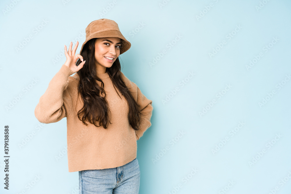 Young indian woman wearing a hat isolated on blue background winks an eye and holds an okay gesture with hand. Young indian woman wearing a hat isolated on blue background winks an eye and holds an okay gesture with hand.