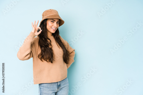 Young indian woman wearing a hat isolated on blue background winks an eye and holds an okay gesture with hand.