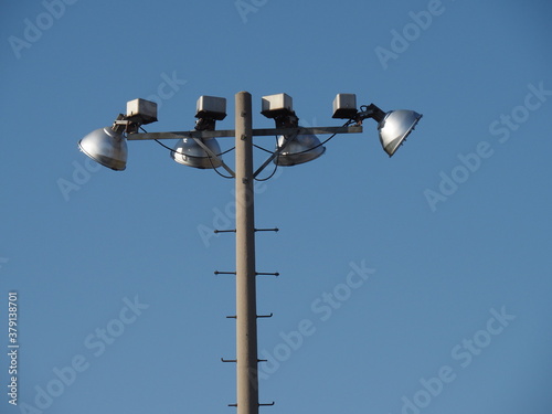 four mercury vapor stadium lights at the top of a concrete pole against a blue sky