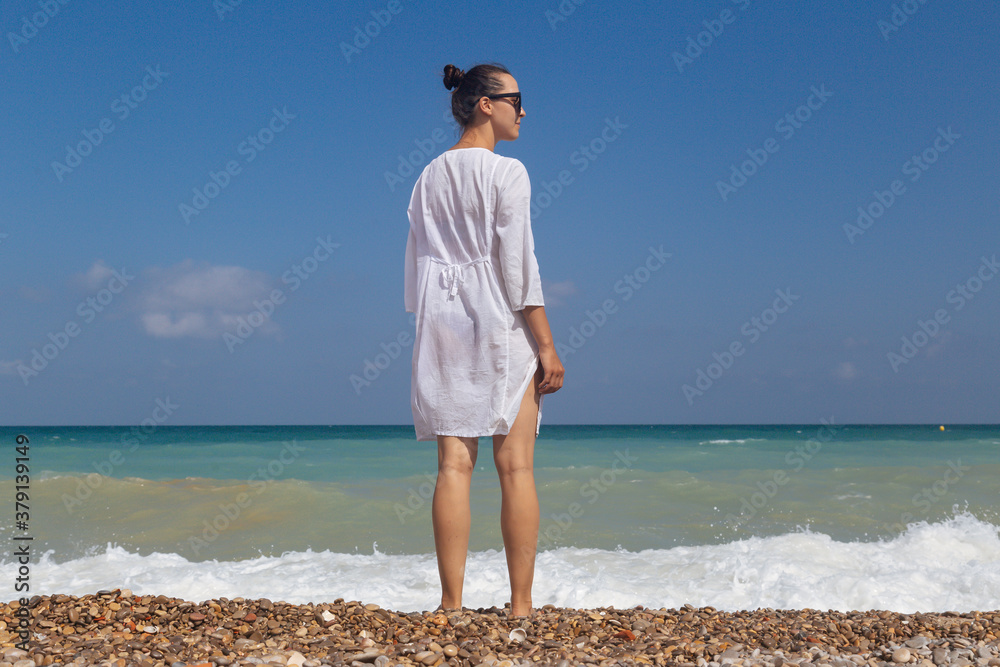 Girl on background of tropical sea at sunset. Woman in summer clothes stands on the beach. Dreaming female. Summer lifestyle, on beach with gazebo and sea in background