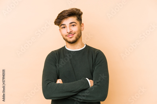 Young arabian man isolated on a beige background who feels confident, crossing arms with determination.